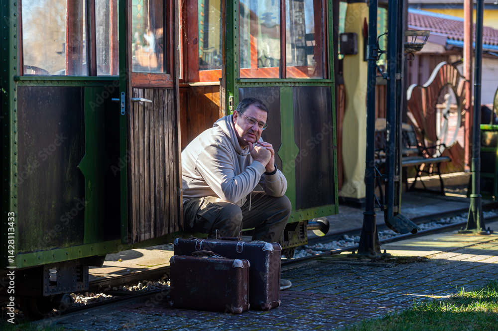 Man sits outside vintage tram station with suitcase on a sunny day