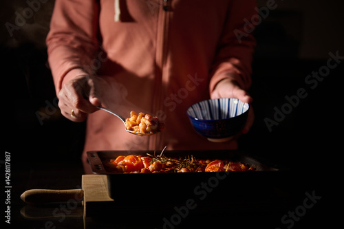 A person serves freshly cooked baked beans garnished with herbs into a blue bowl, captured in a dimly-lit kitchen setting emphasizing deliciousness and care in meal preparation.
