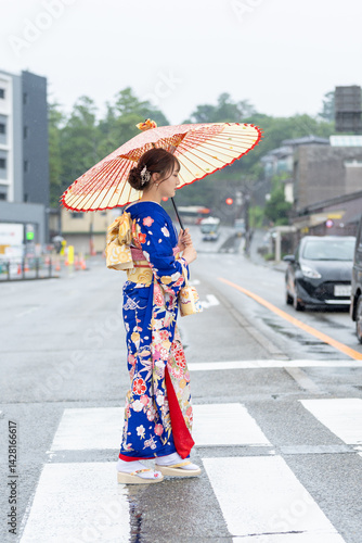 A local city in Japan on a rainy summer day. A Japanese woman wearing a blue teenage kimono holding a Japanese umbrella and walking along a crosswalk.