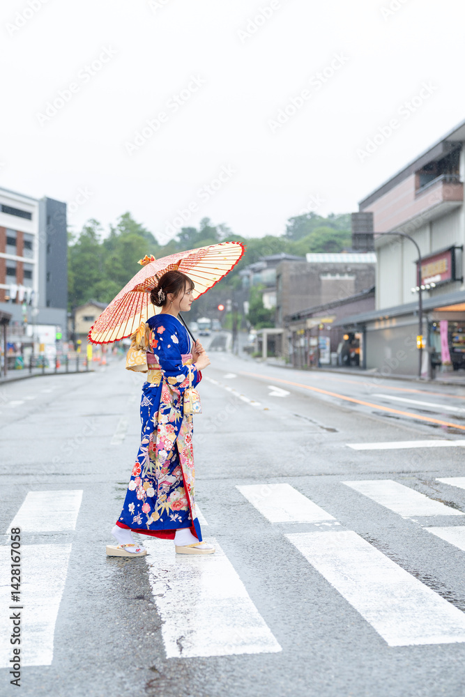 A local city in Japan on a rainy summer day. A Japanese woman wearing a blue teenage kimono holding a Japanese umbrella and walking along a crosswalk.