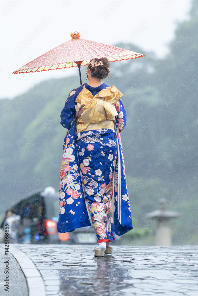 A local city in Japan on a rainy summer day. A Japanese woman wearing a blue teenage kimono holding a Japanese umbrella and climbing a slope. The back view.