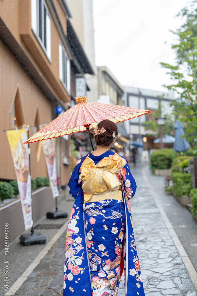 A local city in Japan on a rainy summer day. A Japanese woman wearing a blue teenage kimono holding a Japanese umbrella and climbing a slope. The back view.