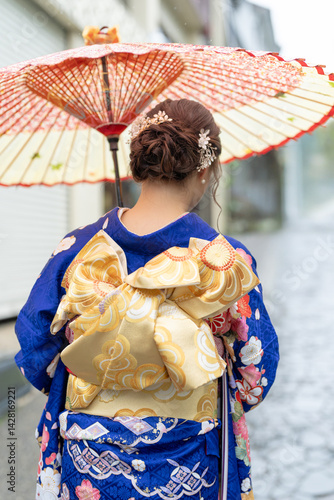 Obraz na płótnie A local city in Japan on a rainy summer day. A Japanese woman wearing a blue teenage kimono holding a Japanese umbrella and climbing a slope. The back view. w salonie