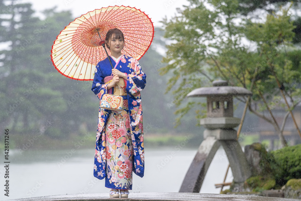 A local city in Japan on a rainy summer day. Japanese woman wearing a blue teenage kimono with a Japanese umbrella standing by a pond in a traditional garden.