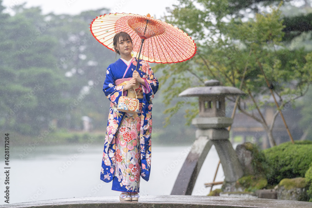 A local city in Japan on a rainy summer day. Japanese woman wearing a blue teenage kimono with a Japanese umbrella standing by a pond in a traditional garden.