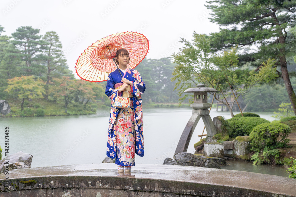 A local city in Japan on a rainy summer day. Japanese woman wearing a blue teenage kimono with a Japanese umbrella standing by a pond in a traditional garden.
