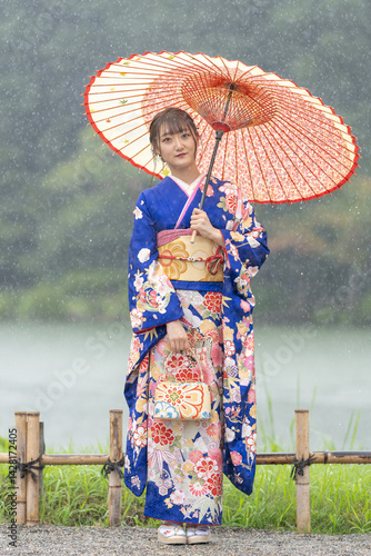 A local city in Japan on a rainy summer day. Japanese woman wearing a blue teenage kimono with a Japanese umbrella standing by a pond in a traditional garden.
