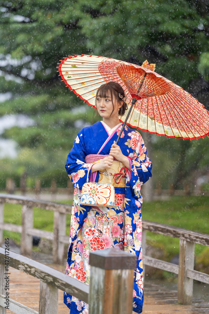 A local city in Japan on a rainy summer day. A Japanese woman wearing a blue teenage kimono holding a Japanese umbrella and walking along a traditional garden bridge.