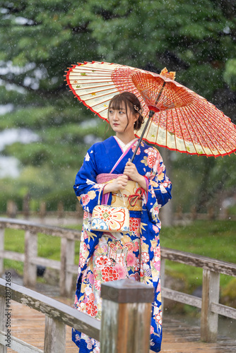 Obraz na płótnie A local city in Japan on a rainy summer day. A Japanese woman wearing a blue teenage kimono holding a Japanese umbrella and walking along a traditional garden bridge. w salonie