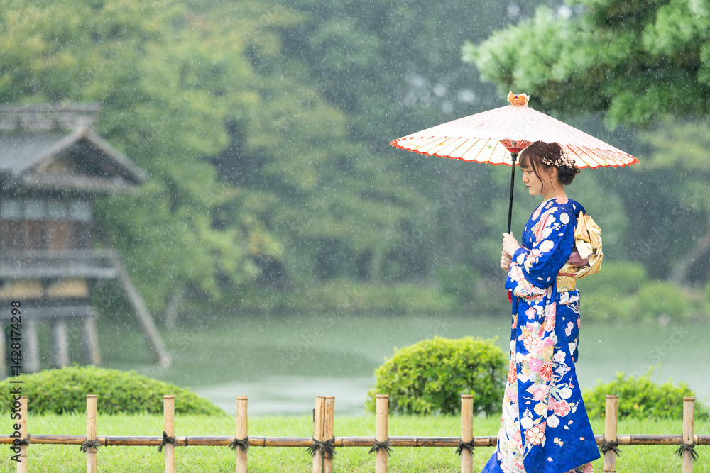 A local city in Japan on a rainy summer day. Japanese woman wearing a blue teenage kimono with a Japanese umbrella walking by a pond in a traditional garden.