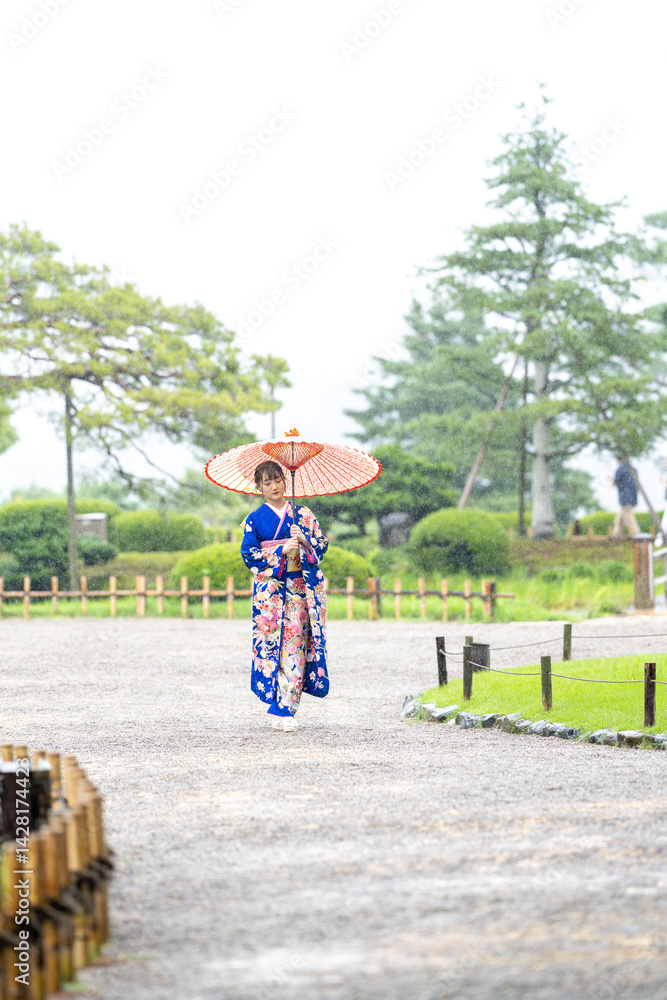 A local city in Japan on a rainy summer day. Japanese woman wearing a blue kimono for teenagers with a Japanese umbrella walking through a traditional garden.