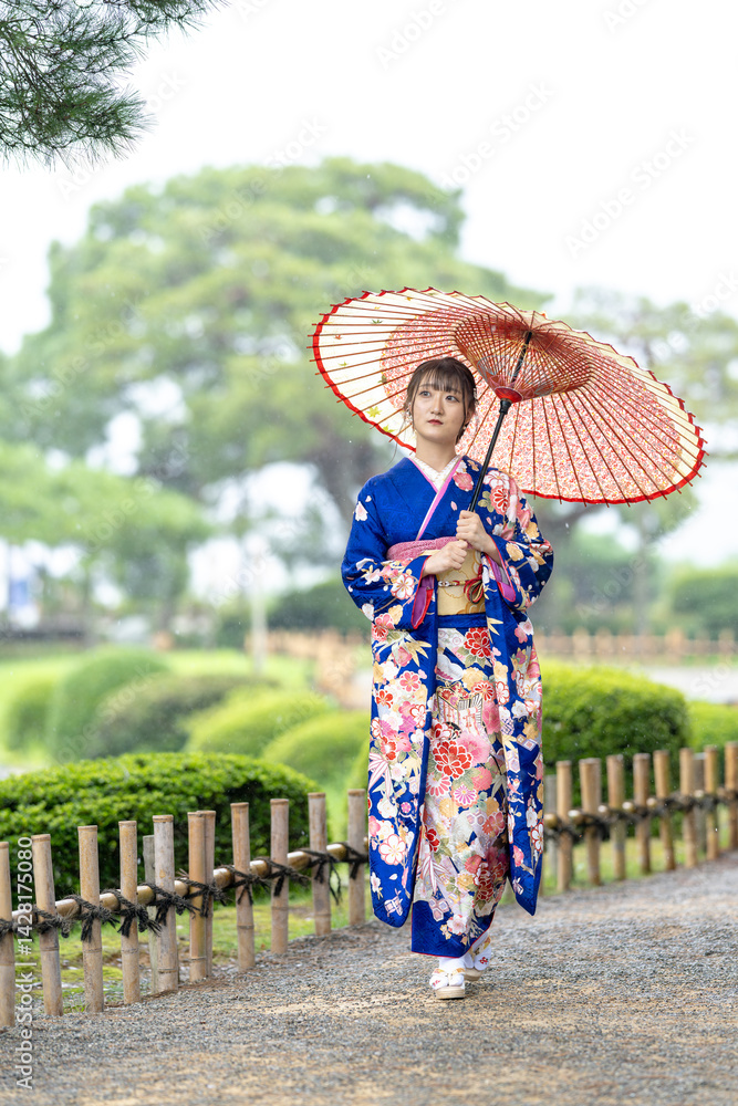 A local city in Japan on a rainy summer day. Japanese woman wearing a blue kimono for teenagers with a Japanese umbrella walking through a traditional garden.