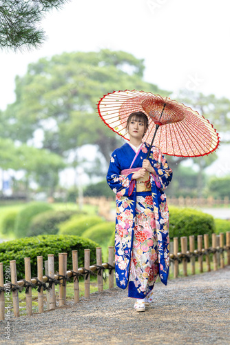Obraz na płótnie A local city in Japan on a rainy summer day. Japanese woman wearing a blue kimono for teenagers with a Japanese umbrella walking through a traditional garden. w salonie