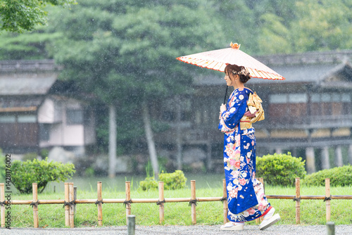 A local city in Japan on a rainy summer day. Japanese woman wearing a blue teenage kimono with a Japanese umbrella walking by a pond in a traditional garden.