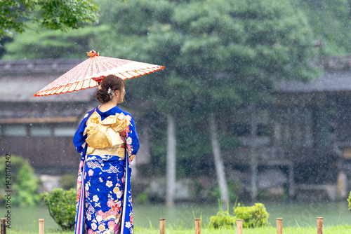 Obraz na płótnie A local city in Japan on a rainy summer day. Japanese woman wearing a blue teenage kimono with a Japanese umbrella standing by a pond in a traditional garden. The back view. w salonie