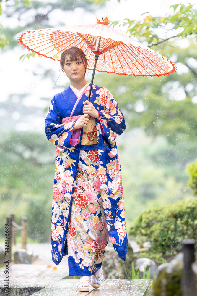 A local city in Japan on a rainy summer day. Japanese woman wearing a blue teenage kimono with a Japanese umbrella walking along a traditional garden bridge.
