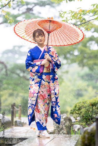A local city in Japan on a rainy summer day. Japanese woman wearing a blue teenage kimono with a Japanese umbrella walking along a traditional garden bridge.