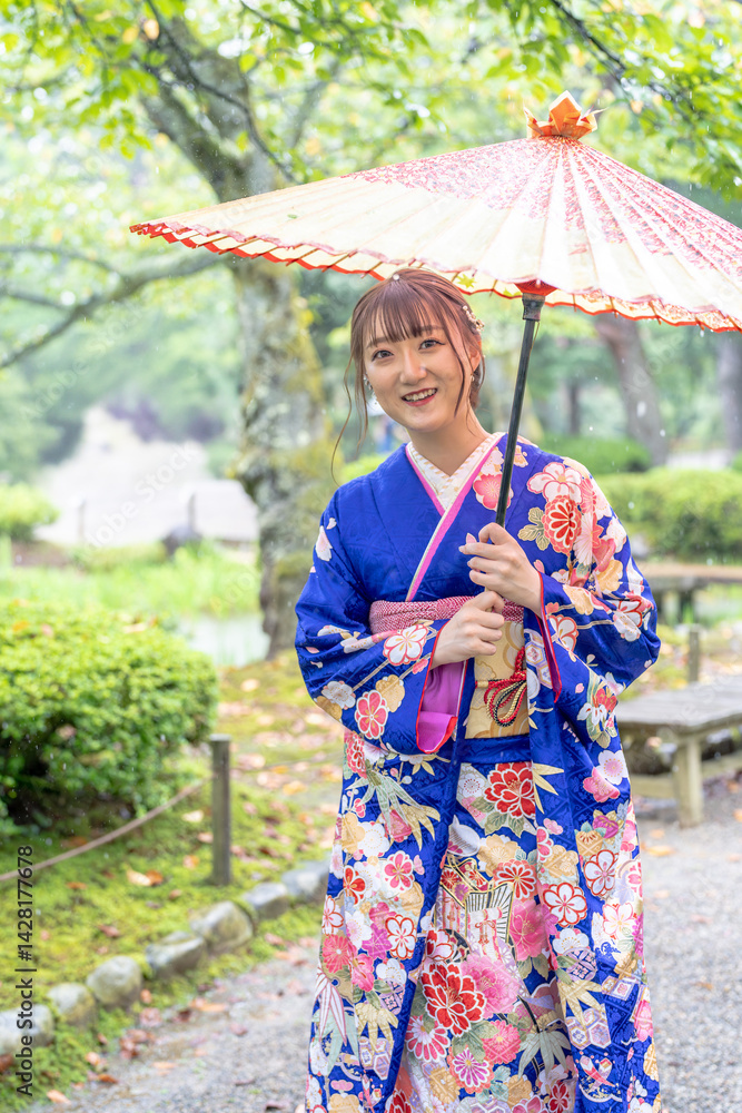 A local city in Japan on a rainy summer day. Japanese woman wearing a blue kimono for teenagers with a Japanese umbrella walking through a traditional garden.