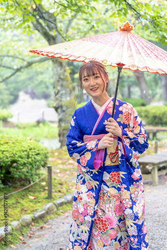 Obraz na płótnie A local city in Japan on a rainy summer day. Japanese woman wearing a blue kimono for teenagers with a Japanese umbrella walking through a traditional garden. w salonie