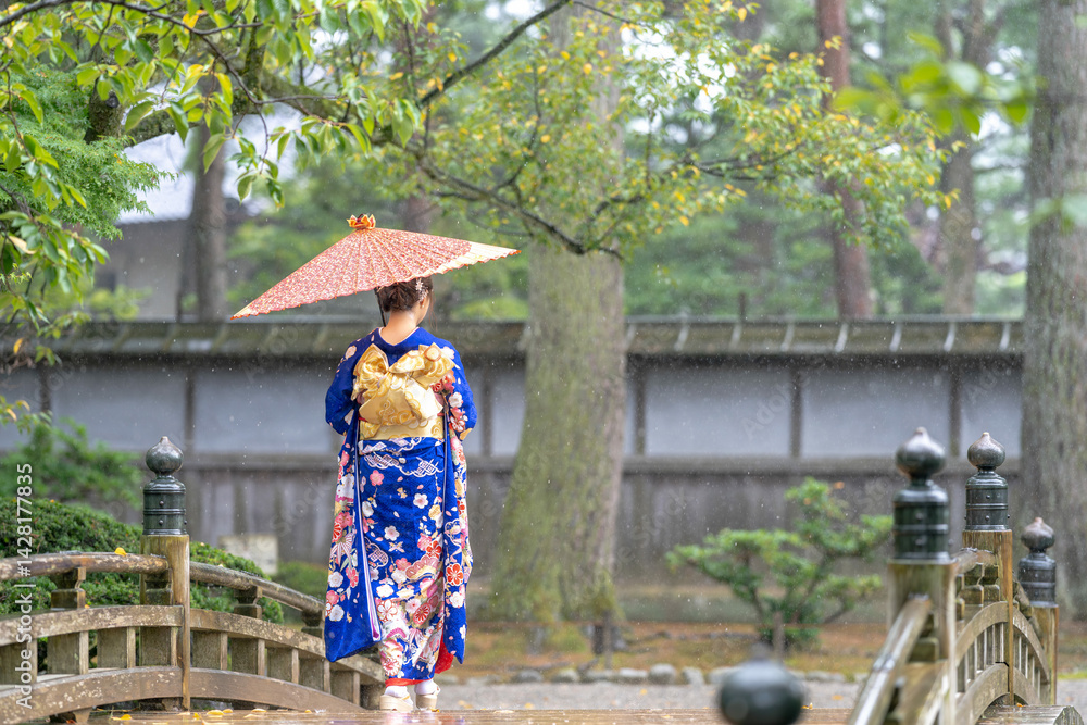 A local city in Japan on a rainy summer day. Japanese woman wearing a blue teenage kimono with a Japanese umbrella walking along a traditional garden bridge.