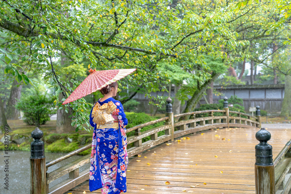 A local city in Japan on a rainy summer day. Japanese woman wearing a blue teenage kimono with a Japanese umbrella walking along a traditional garden bridge.