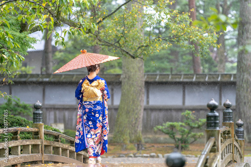 Obraz na płótnie A local city in Japan on a rainy summer day. Japanese woman wearing a blue teenage kimono with a Japanese umbrella walking along a traditional garden bridge. w salonie