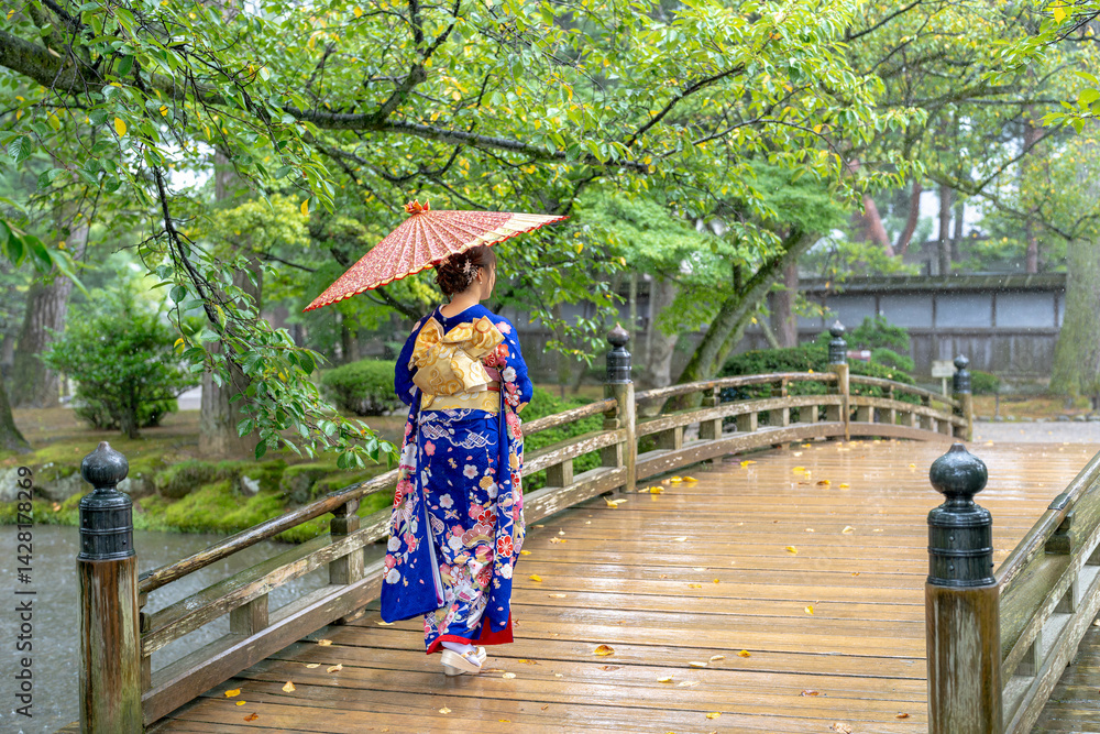 A local city in Japan on a rainy summer day. Japanese woman wearing a blue teenage kimono with a Japanese umbrella walking along a traditional garden bridge.