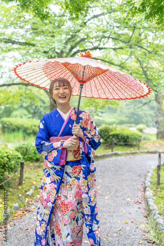 Obraz na płótnie A local city in Japan on a rainy summer day. Japanese woman wearing a blue kimono for teenagers with a Japanese umbrella walking through a traditional garden. w salonie
