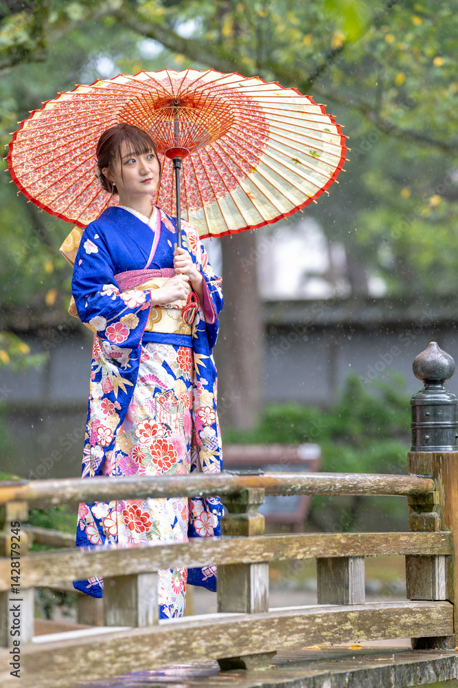 A local city in Japan on a rainy summer day. Japanese woman wearing a blue teenage kimono with a Japanese umbrella standing on a traditional garden bridge.