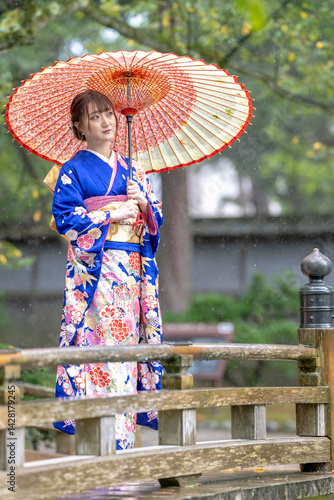 Obraz na płótnie A local city in Japan on a rainy summer day. Japanese woman wearing a blue teenage kimono with a Japanese umbrella standing on a traditional garden bridge. w salonie