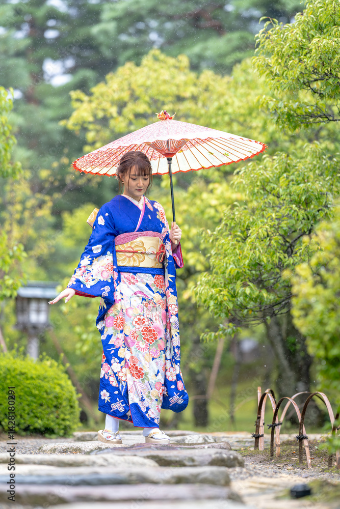 A local city in Japan on a rainy summer day. Japanese woman wearing a blue kimono for teenagers with a Japanese umbrella walking through a traditional garden.