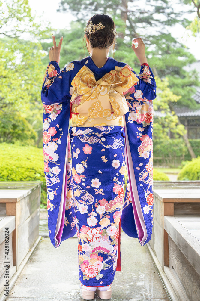 A local city in Japan on a rainy summer day. A Japanese woman wearing a blue teenage kimono in a traditional garden. She expresses 2 and 0 with her hands. The back view.