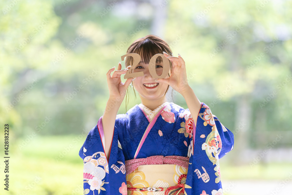A local city in Japan on a rainy summer day. A Japanese woman wearing a blue teenage kimono in a traditional garden. She holds blocks of 2 and 0 in her hand and laughs. Celebration of 20 years old.