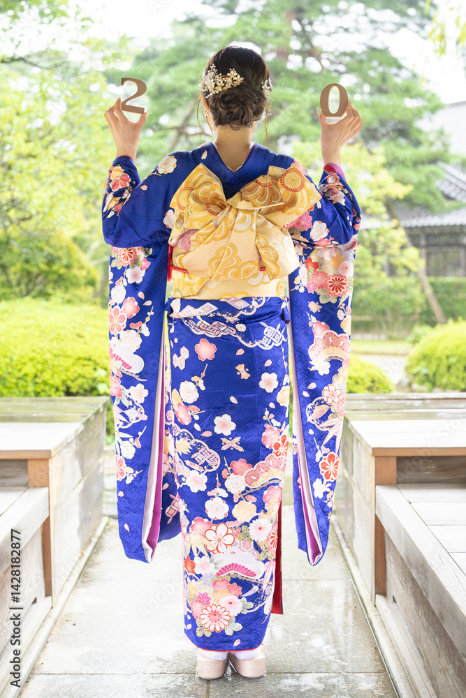 A local city in Japan on a rainy summer day. A Japanese woman wearing a blue teenage kimono in a traditional garden. She holds blocks of 2 and 0 in her hand. The back view.