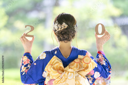 A local city in Japan on a rainy summer day. A Japanese woman wearing a blue teenage kimono in a traditional garden. She holds blocks of 2 and 0 in her hand. The back view.