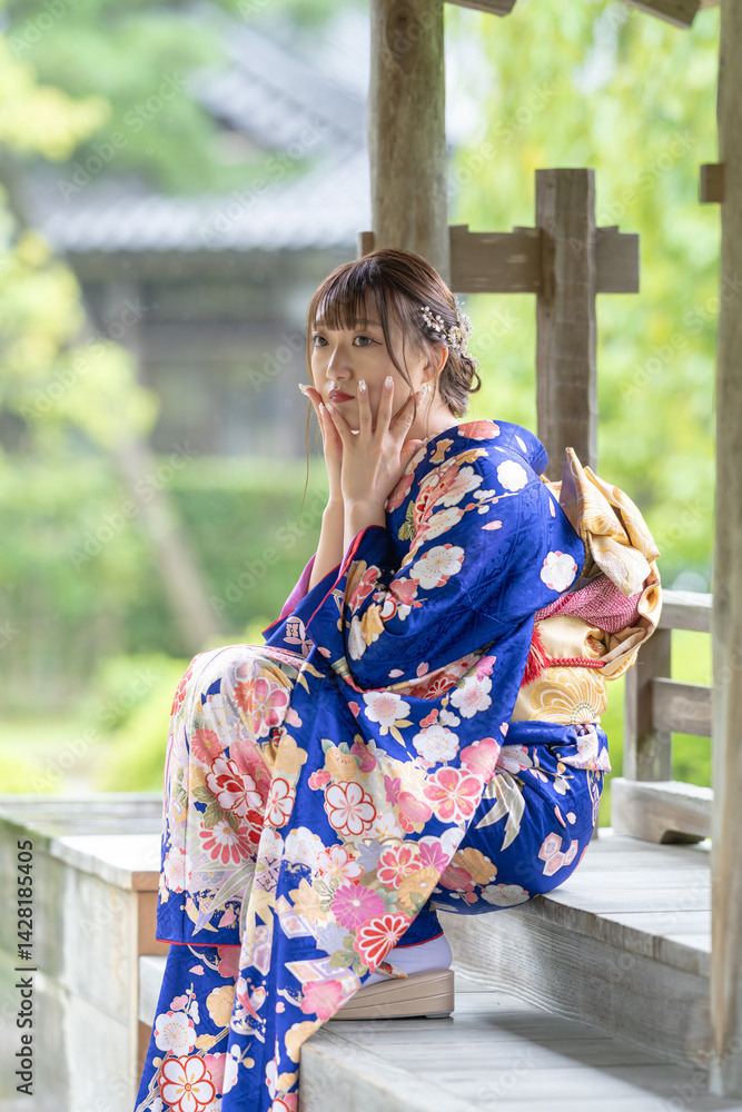 A local city in Japan on a rainy summer day. A Japanese woman wearing a blue teenage kimono in a traditional garden. She is sitting on a wooden chair. She puts her hand on her cheek.