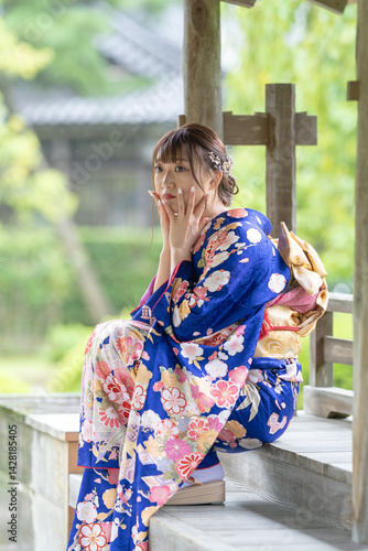 Obraz na płótnie A local city in Japan on a rainy summer day. A Japanese woman wearing a blue teenage kimono in a traditional garden. She is sitting on a wooden chair. She puts her hand on her cheek. w salonie