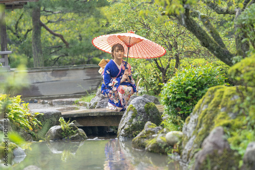 A local city in Japan on a rainy summer day. A Japanese woman wearing a blue teenage kimono in a traditional garden. She holds an umbrella and sits looking out at the river.