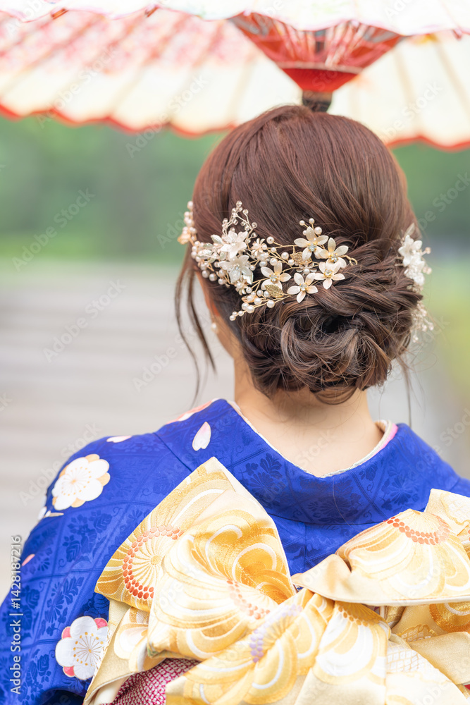 A local city in Japan on a rainy summer day. Japanese woman wearing a blue kimono for teenagers with a Japanese umbrella walking through a traditional garden. The back view.