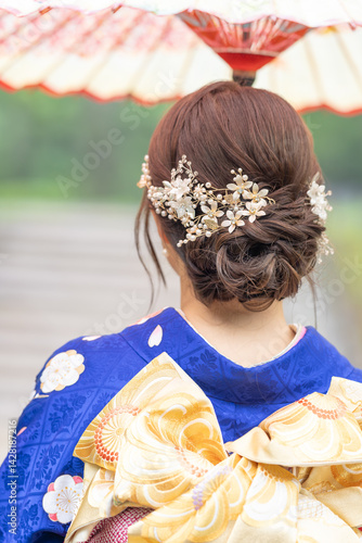 Obraz na płótnie A local city in Japan on a rainy summer day. Japanese woman wearing a blue kimono for teenagers with a Japanese umbrella walking through a traditional garden. The back view. w salonie