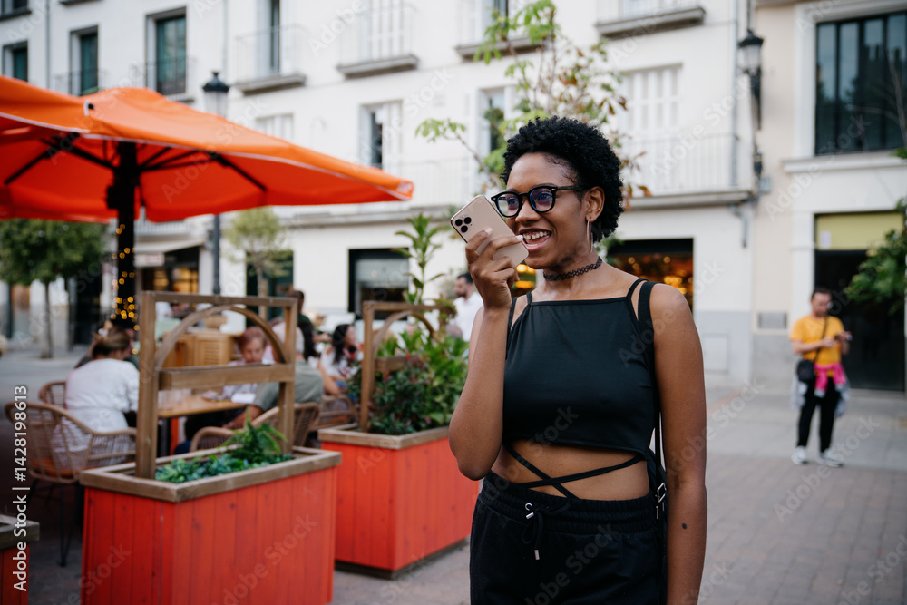 Happy young African American woman sending voice message outdoors, smiling while using smartphone on lively city street with orange umbrellas and outdoor restaurant in background
