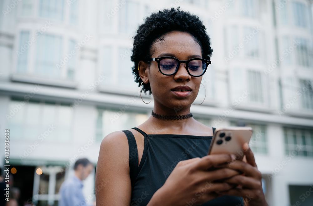 Close-up of stylish young black woman using smartphone outdoors, reading screen with focus, wearing glasses and black choker in modern urban environment