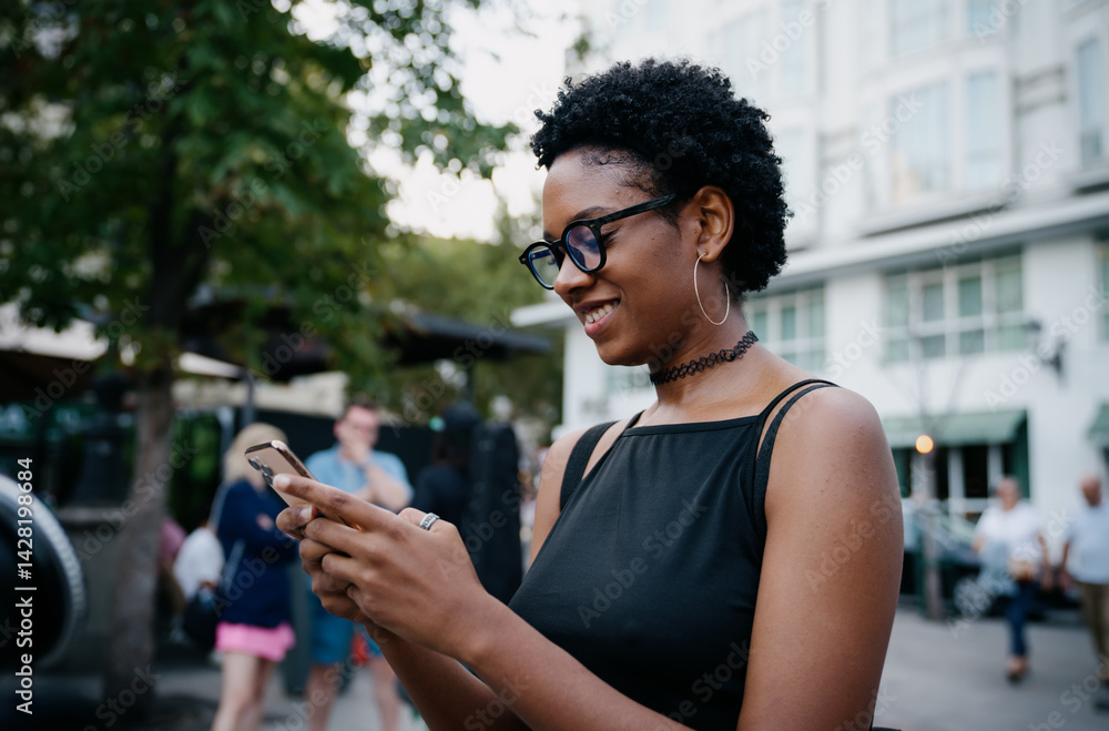 Smiling young black woman interacting with smartphone outdoors, wearing glasses and hoop earrings, enjoying digital communication in casual modern street environment