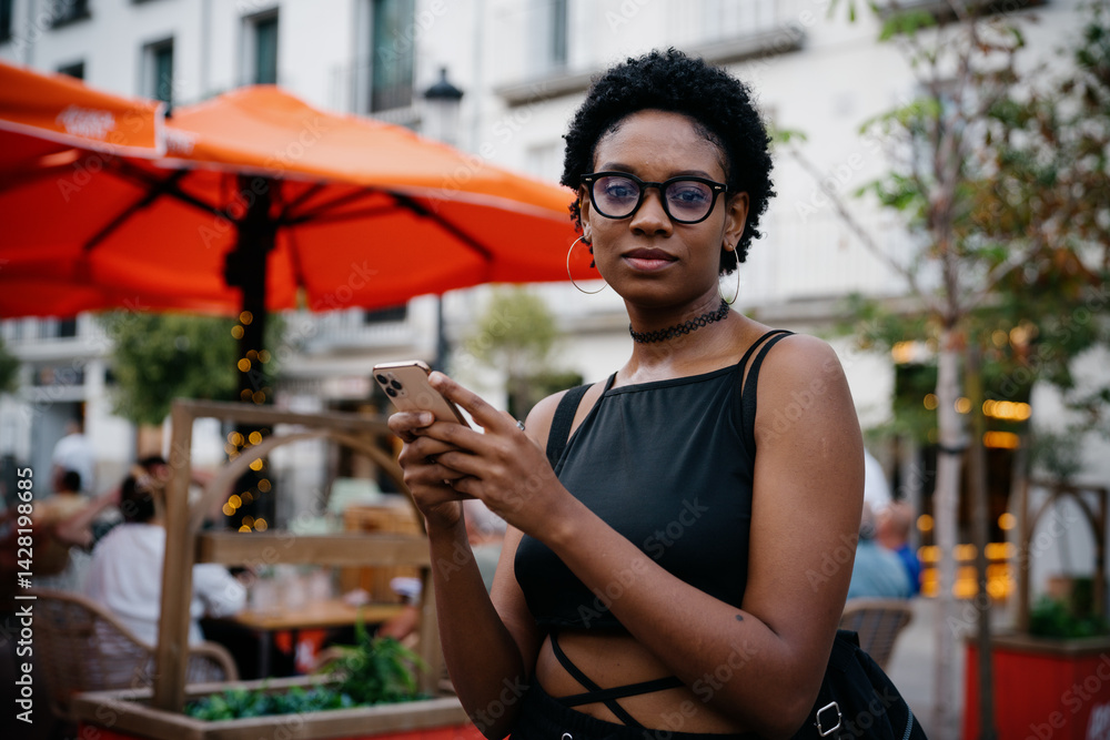 Confident young African American woman holding smartphone with serious expression, standing on city street near outdoor café with orange parasols during summer afternoon