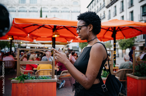 Obraz na płótnie Young African American woman using smartphone while walking past outdoor café with vibrant orange umbrellas, dressed in black outfit, focused on mobile screen in urban summer setting w salonie