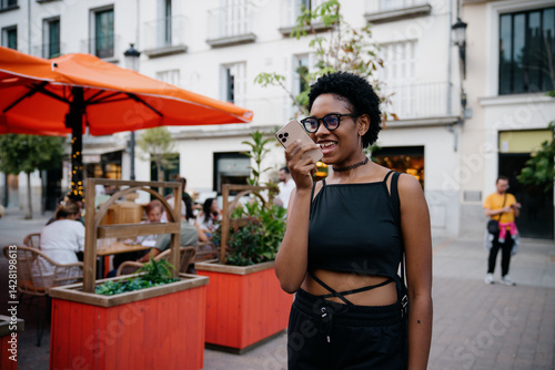 Obraz na płótnie Happy young African American woman sending voice message outdoors, smiling while using smartphone on lively city street with orange umbrellas and outdoor restaurant in background w salonie