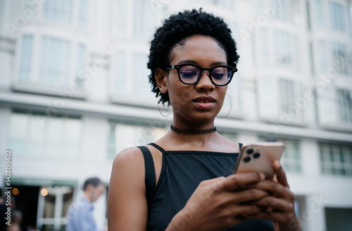 Obraz na płótnie Close-up of stylish young black woman using smartphone outdoors, reading screen with focus, wearing glasses and black choker in modern urban environment w salonie