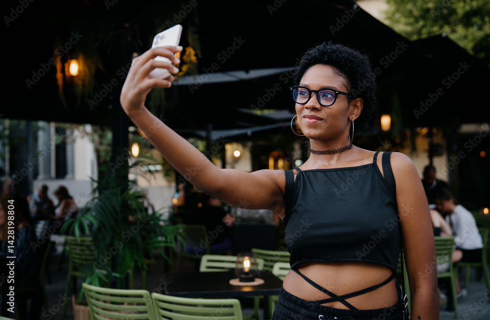 Happy young woman in stylish outfit smiles as she records vlog segment or chats on video call, standing near café tables under cozy lights in relaxed urban setting