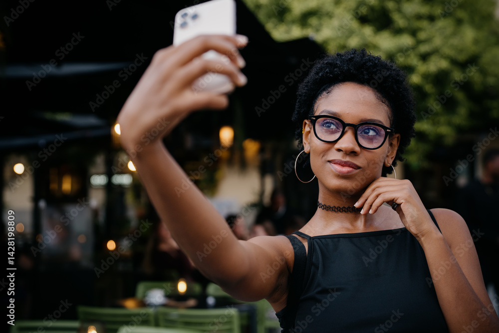 Confident millennial woman poses for a cute selfie at cozy café, holding her phone high and smiling slightly, creating personal content for her social feed or blog audience