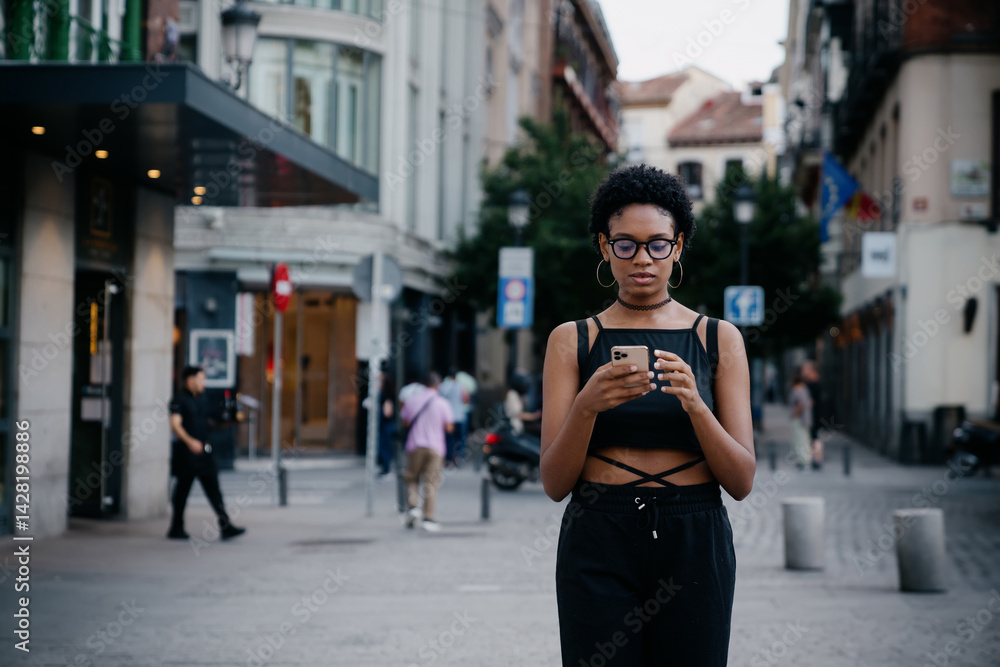 Young Black woman walks through city street while browsing smartphone, focused on playlist, podcast, or audiobook, merging tech use with an urban lifestyle and casual style